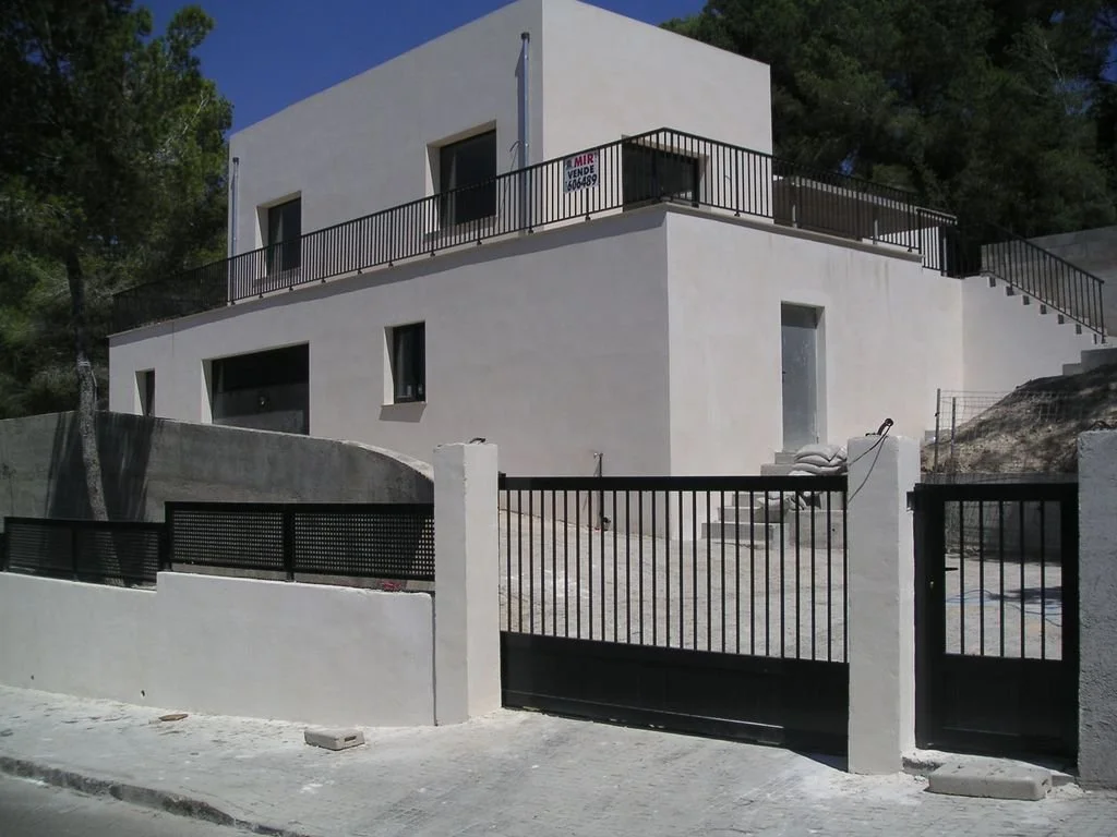 Casa moderna de dos niveles con paredes blancas, barandales negros y un portón en la entrada, en un área rodeada de árboles.