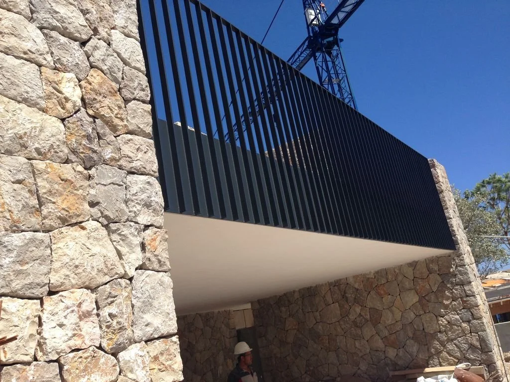 Estructura de edificio en construcción con muro de piedra y barandilla negra, con una grúa en el fondo, cielo azul y árbol a un lado.