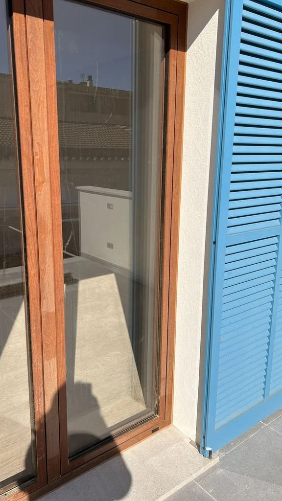 Puerta de vidrio con marco de madera, parte de una fachada con muro blanco y una tienda con persianas azules.