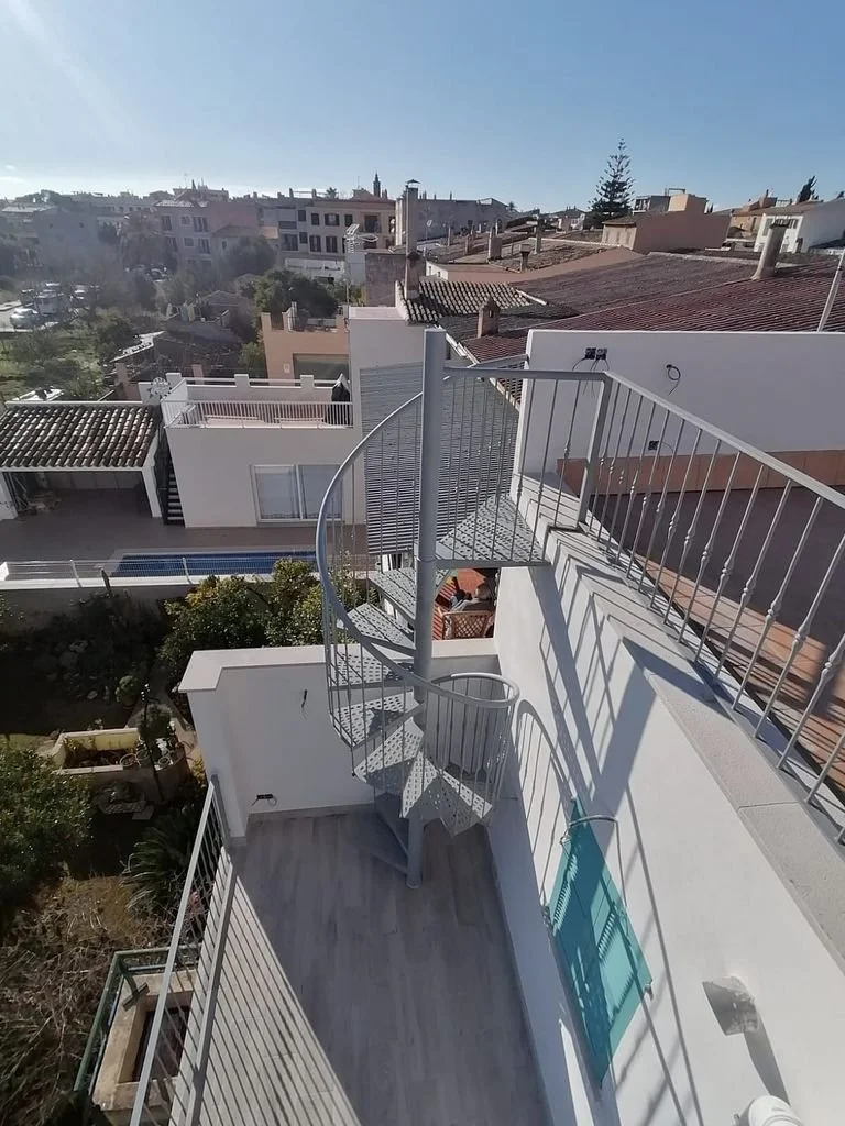 Vista desde un balcón en la azotea con escalera de caracol de metal, barandal, piso de madera y vistas de edificios residenciales y tejados en una ciudad bajo cielo despejado.