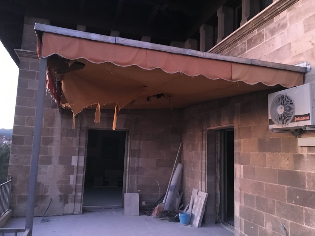 Terraza exterior con toldo desgastado y cojín rasgado, pared de ladrillos, ventilador de aire acondicionado y materiales de construcción junto a la pared.
