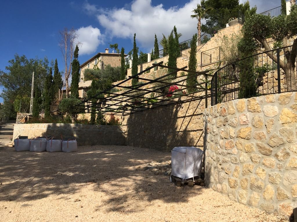 Jardín o patio con estructura metálica en sombra, mas plantas y árboles en un nivel superior, paredes de piedra, y bolsas de arena o materiales de jardinería en el suelo.
