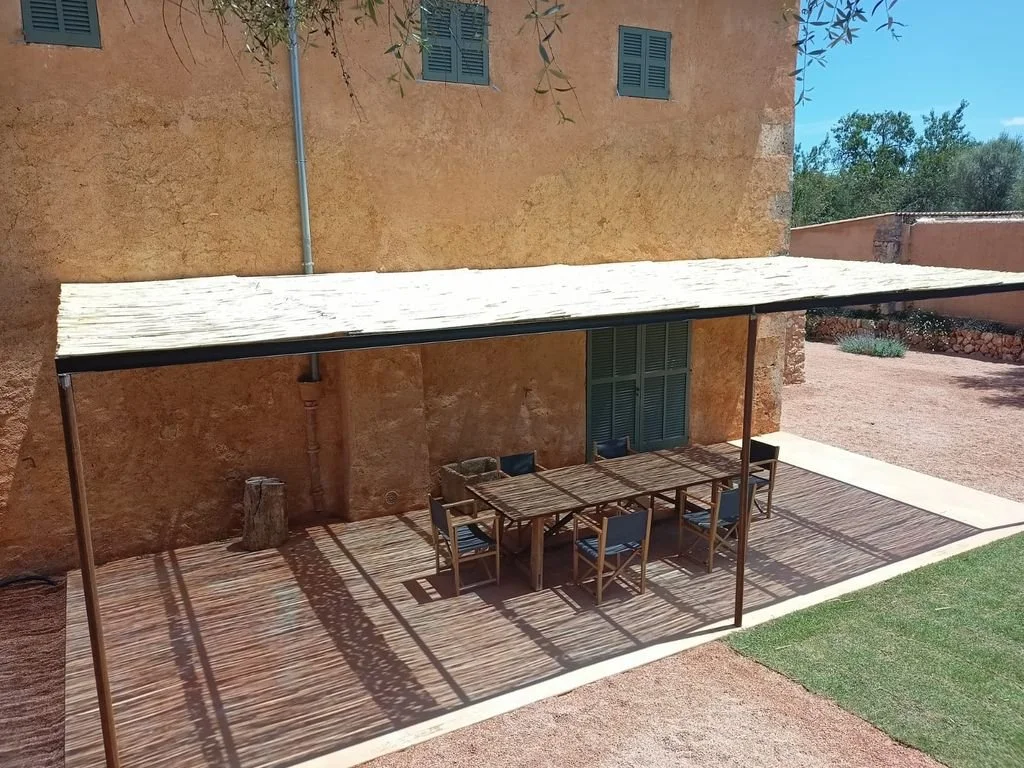 Terraza exterior con mesa de madera y sillas, sombra de toldo de tela sobre la superficie y la pared de fondo es de color terracota.