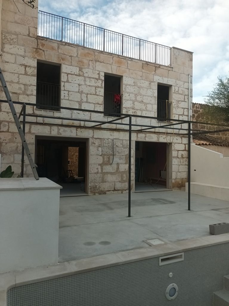 Edificio en construcción con paredes de piedra y terraza en la parte superior, estructura de metal en el patio y piscina en primer plano.