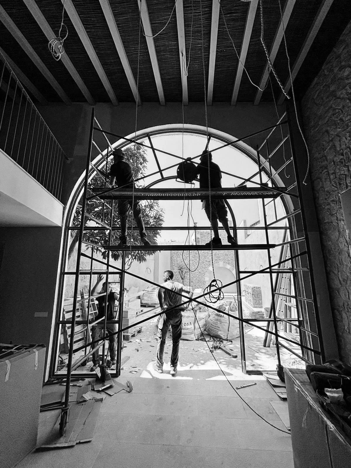 Cuatro trabajadores de construcción en un andamio dentro de un edificio en construcción. Están trabajando en la entrada principal, que tiene un arco grande de cristal. El área está en fase de acabado, con materiales de construcción dispersos alrededor.
