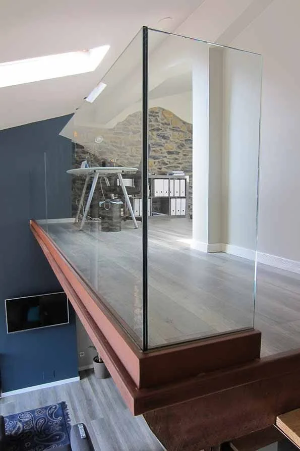 Vista de un rincón de un apartamento con una pared de roca expuesta, un escritorio en la esquina, y un espacio vacío con suelo de madera, rodeado por un muro de cristal que forma una especie de balcón o separación interior.