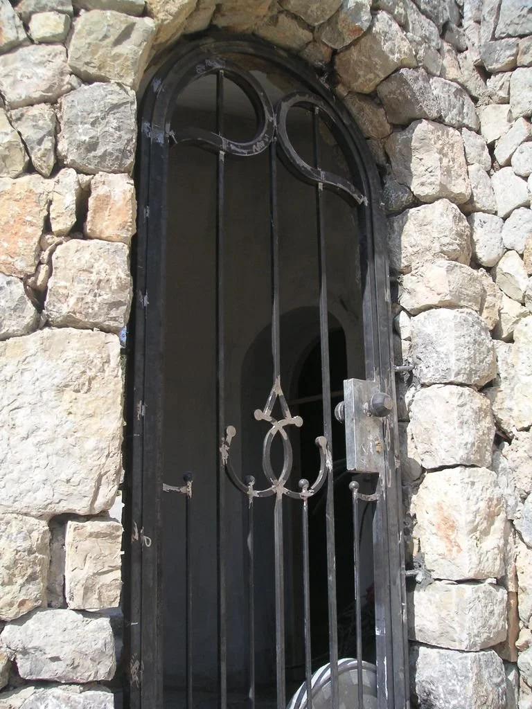 Puerta de hierro con reja decorativa, ubicada en marco de piedra en una pared de piedra.
