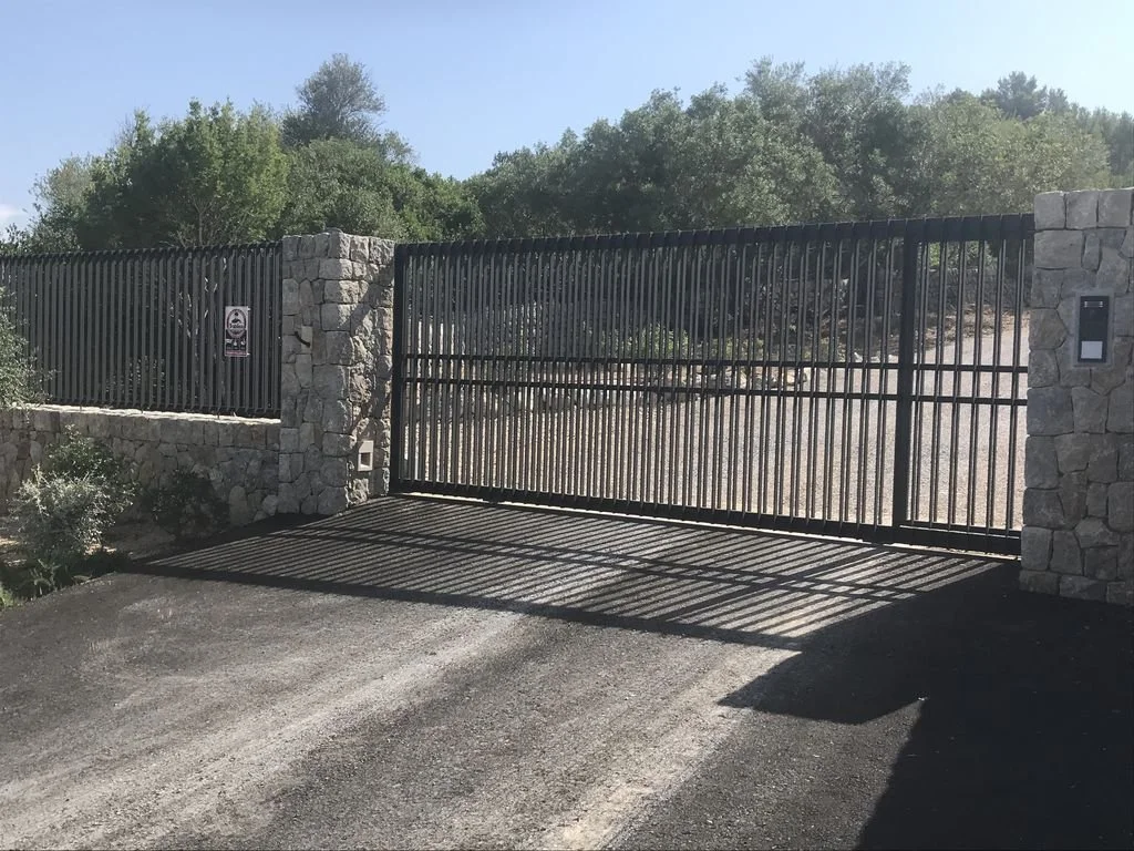 Puerta automática de metal con reja en entrada de finca, con muros de piedra a los lados y vegetación densa de árboles en el fondo.