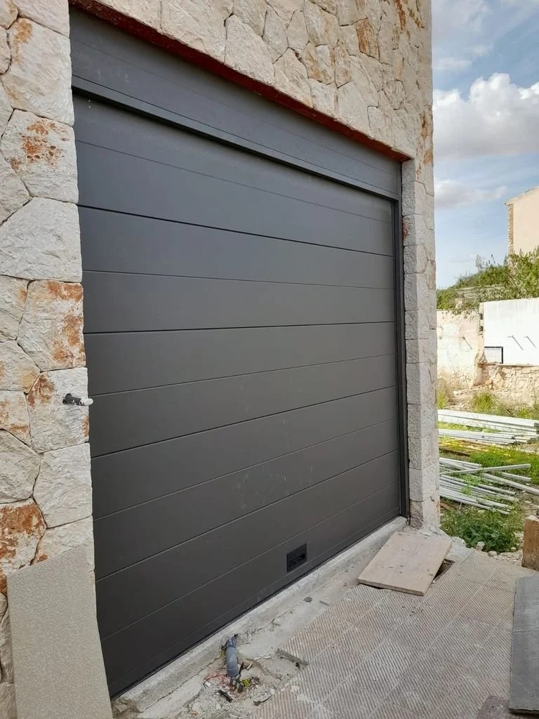 Una puerta de garaje moderna de color gris oscuro instalada en una estructura de piedra, con un entorno exterior en construcción.
