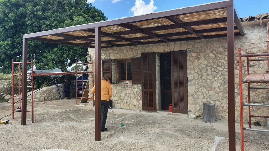 Una persona barriendo frente a una casa con paredes de piedra y ventanas con persianas de madera, y un techo en construcción en un patio pavimentado.