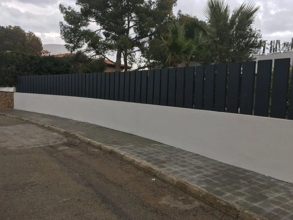 Muro de cemento blanco y cerca de lámina negra en una calle con acera de piedra, árboles y vegetación al fondo.