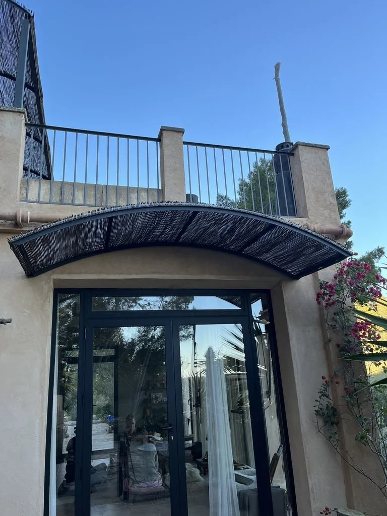 Vista de la fachada de una casa con puertas de cristal, un balcón con barandilla y una estructura de adorno en la parte superior, rodeada de plantas y flores.