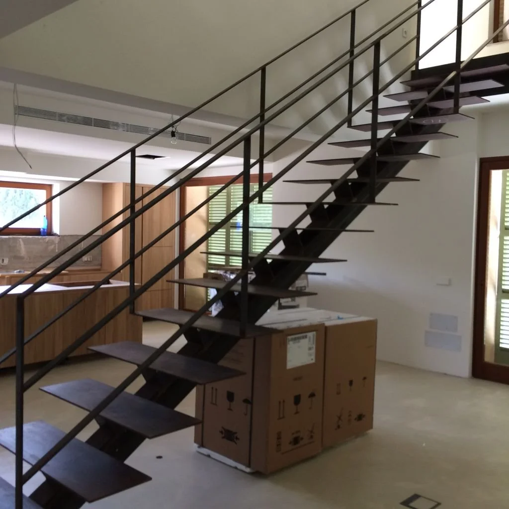 Escalera de madera y metal en una sala de vivienda en construcción, con cajas en el suelo y ventanas con persianas verdes.
