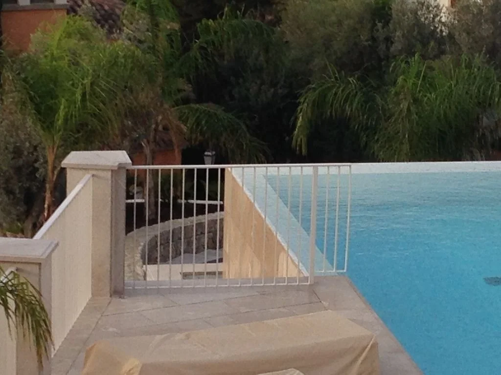 Piscina con vista a un área con plantas y árboles, cerca de una cerca blanca.
