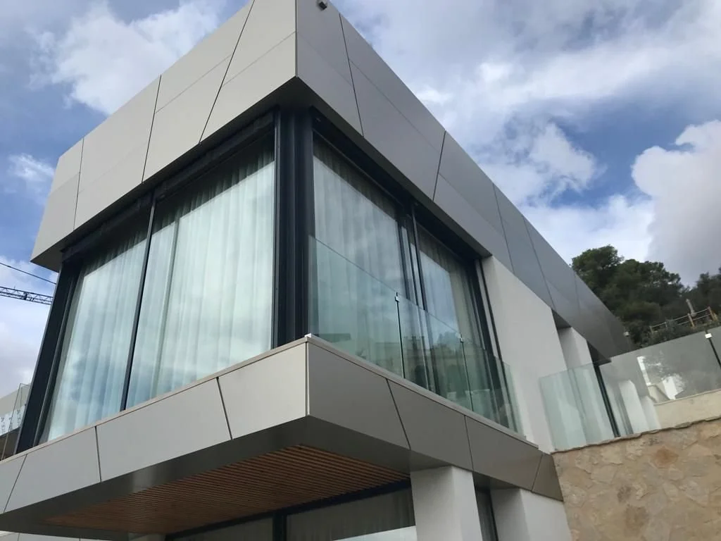Edificio moderno de dos pisos con paredes de vidrio y acabado en colores gris y blanco, con cielo nublado de fondo.