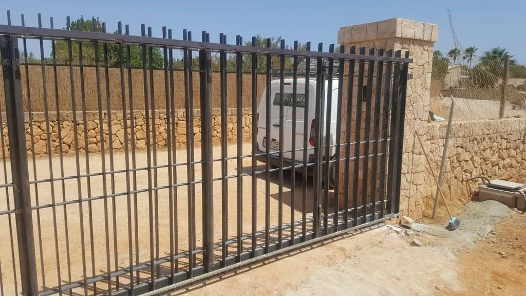 Puerta de hierro con rejas, muro de piedra, auto blanco estacionado detrás de la puerta, en un área de tierra, en un entorno con muro de piedra y estructura de aluminio.