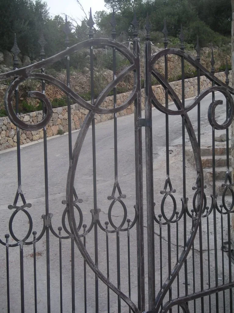 Puerta de hierro con forma de corazón decorativa, en un camino de piedra y vegetación en el fondo.