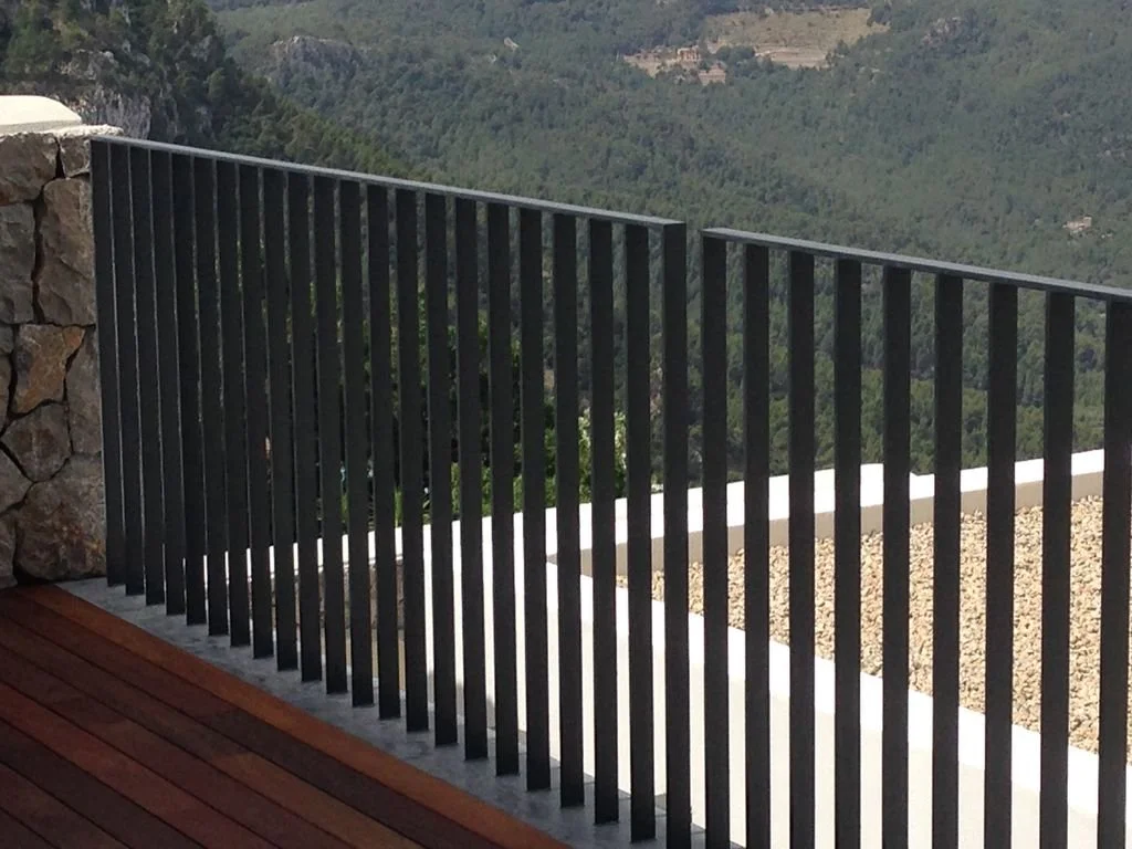 Barandal negro en un balcón de madera, con vista a un paisaje montañoso con árboles.