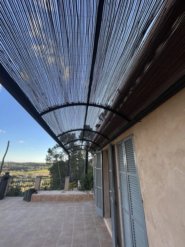 Vista de un balcón con techo de cañizo de madera, puertas de madera pintadas de gris, y panorama de naturaleza con árboles y cielo azul en el fondo
