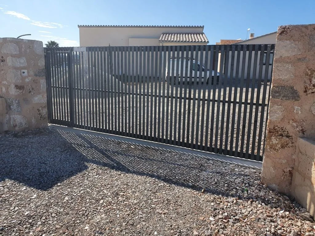 Puerta de metal con listones verticales, instalada en un marco, que da acceso a un patio con grava y vista a casas y un auto en un fondo con cielo despejado.