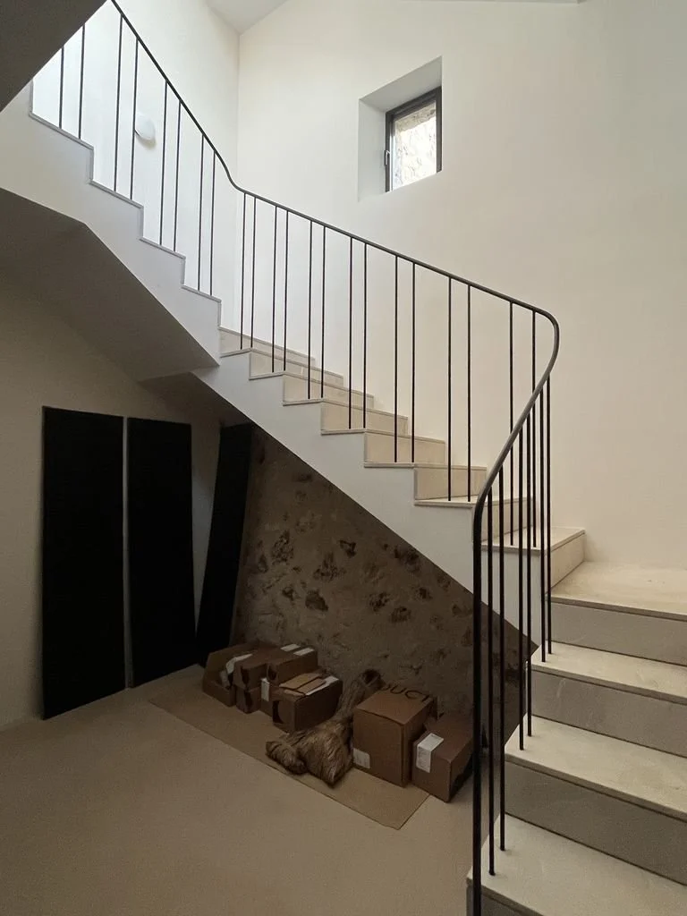 Escalera interior de una casa con paredes blancas, ventanas pequeñas, pisos de piedra y varias cajas apiladas en la esquina.