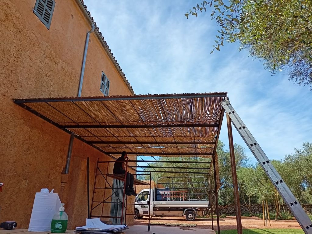 Persona instalando una estructura de sombra de cañas o madera en un área exterior con camino de tierra, junto a una pared de casa con paredes color terracota y ventanas de rejas. En el fondo hay árboles y un cielo despejado.