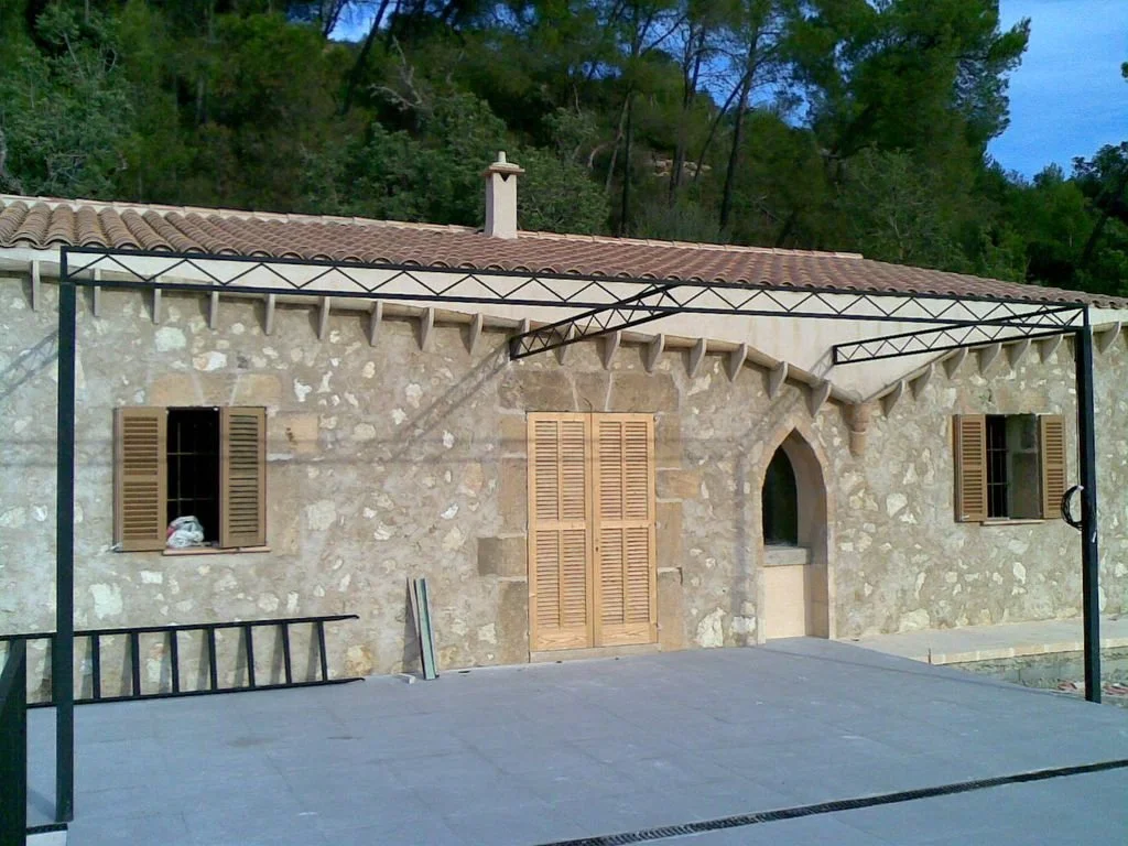 Casa de piedra con ventanas con persianas de madera y puerta en medio, en un entorno natural con árboles, vista de la terraza en primer plano.