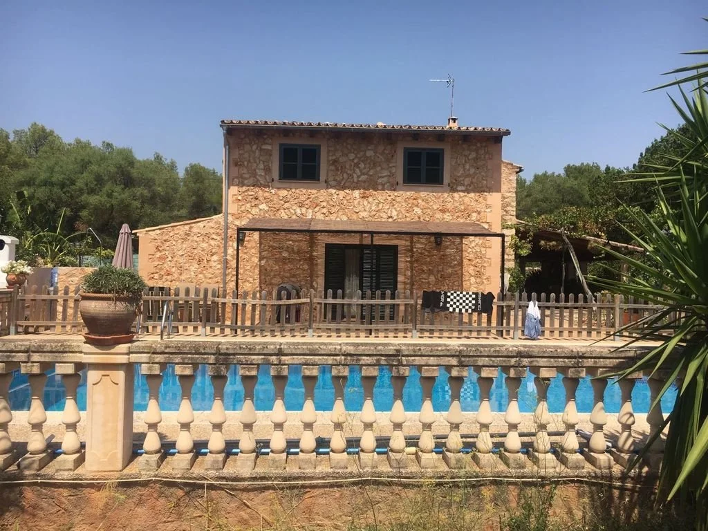 Casa de piedra con dos pisos y ventanas pequeñas, rodeada de árboles, con una piscina en primer plano y una cerca de balaustres, bajo un cielo despejado.