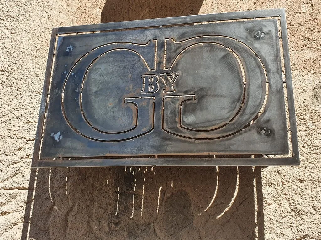 Placa de metal con las letras 'G' y 'O' y la palabra 'BY' en medio, colocada en una superficie de tierra. 