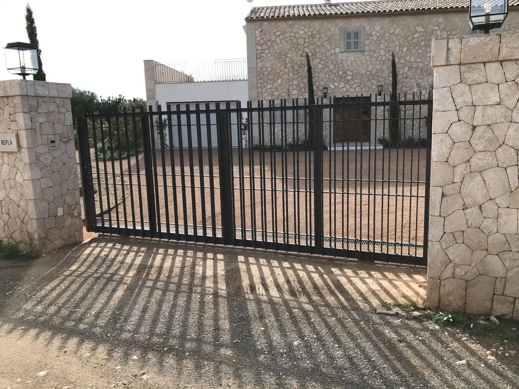 Puerta de rejas de metal negro que cierra un ingreso a un patio de casa, con muros de piedra en los lados y sombra de las rejas en el suelo.