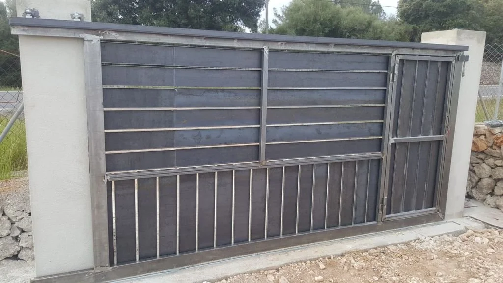 Puerta metálica enrejada instalada en muro de cemento, con fondo de área con césped y árboles.