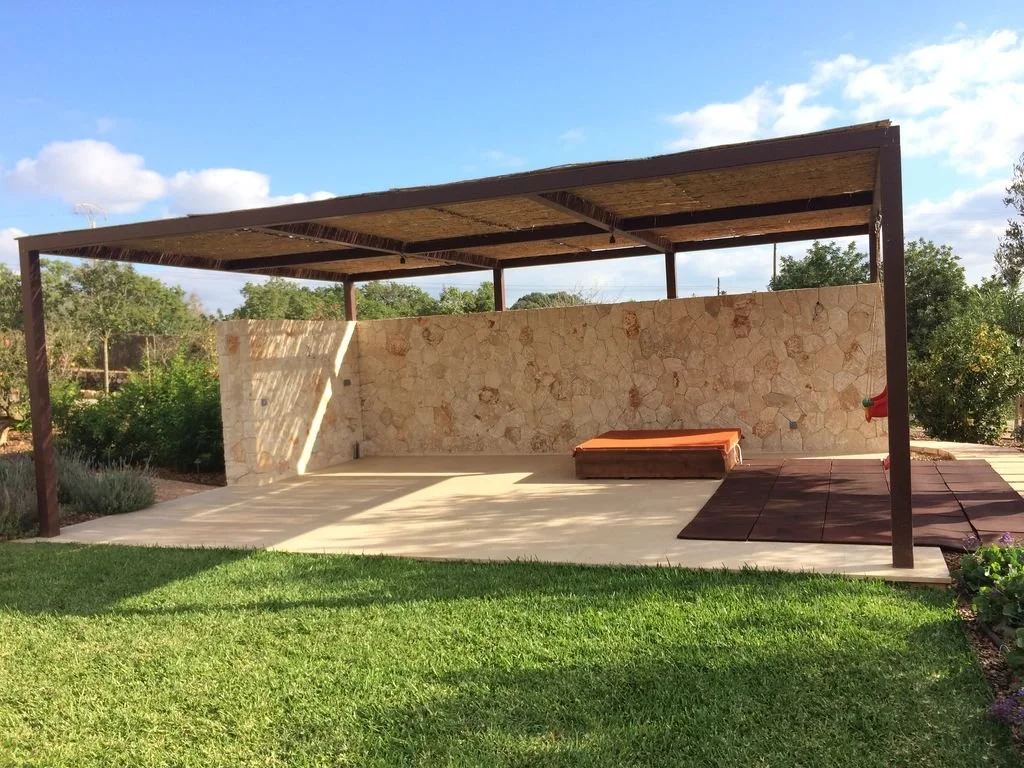 Estructura al aire libre con techo de madera, muro de piedra, áreas de césped y pavimento de concreto en un jardín.