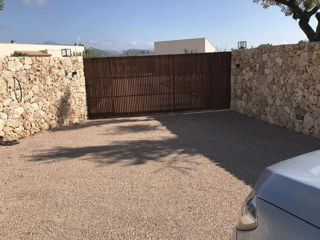 Entrada a un garaje o patio cerrado con muro de piedra y puerta de madera, hay sombra de árboles y parte de un coche en la esquina inferior derecha.