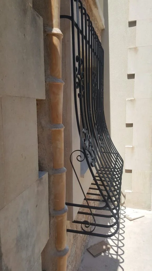 Barandillas de hierro negro adornadas con detalles en espiral, instaladas en una fachada de piedra beige con tuberías de PVC en la esquina izquierda, en una fachada de edificio.