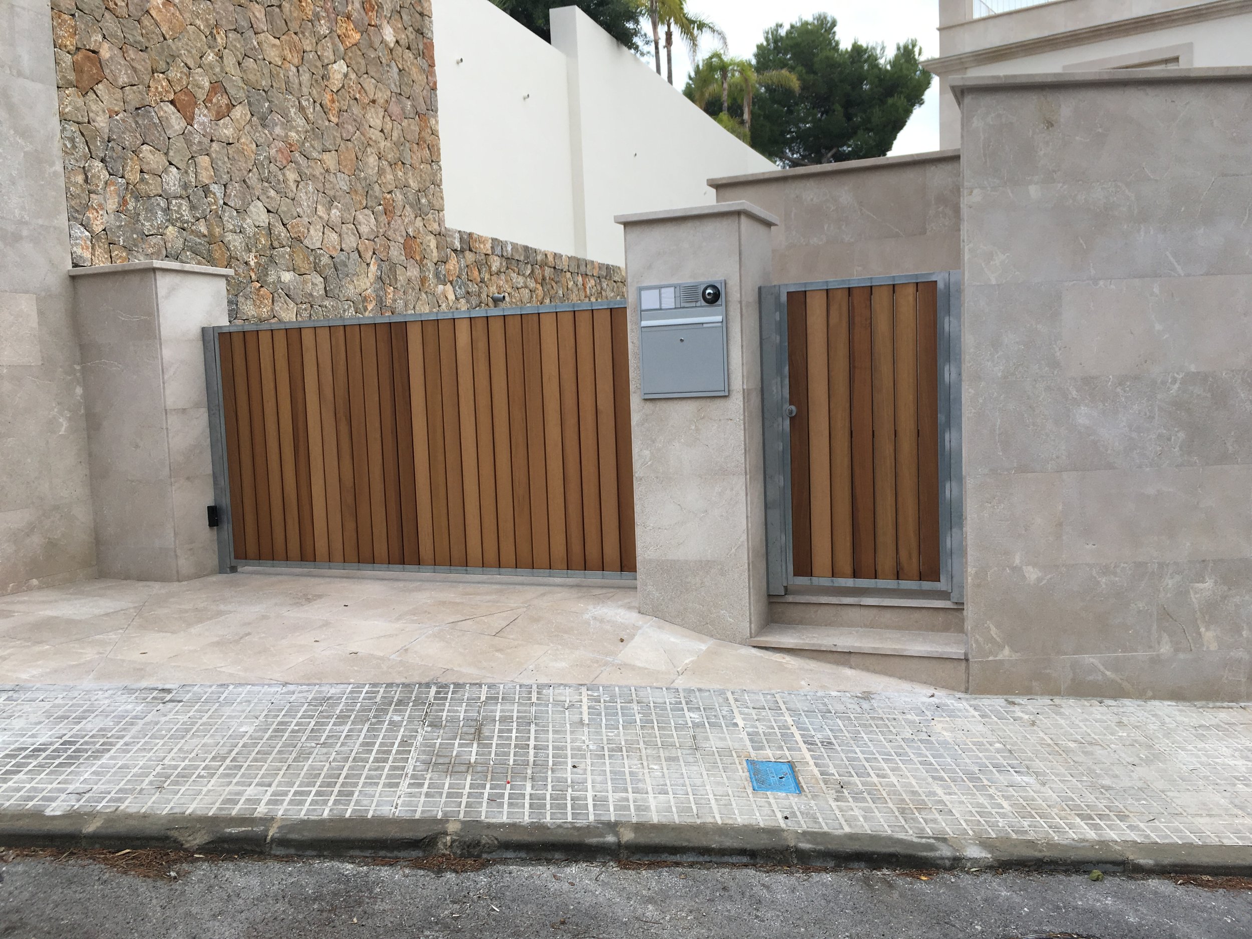 Vista de una entrada de una propiedad con muro de piedra decorativa, puerta de madera con marco metálico, buzón y caseta de vigilancia o control de acceso, con aceras y calle en primer plano y árboles verdes al fondo.