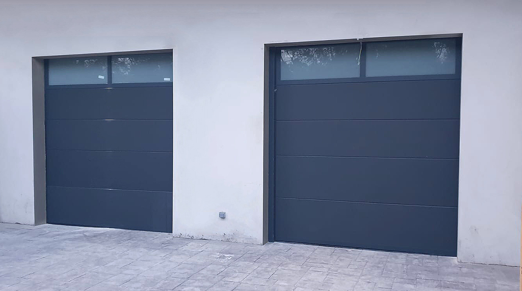 Dos puertas de garaje modernas de color gris oscuro en una pared blanca, con ventanas rectangulares en la parte superior.
