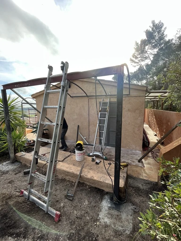 Persona trabajando en la estructura de una construcción o reparación en un área exterior, con escaleras, manguera y herramientas alrededor.