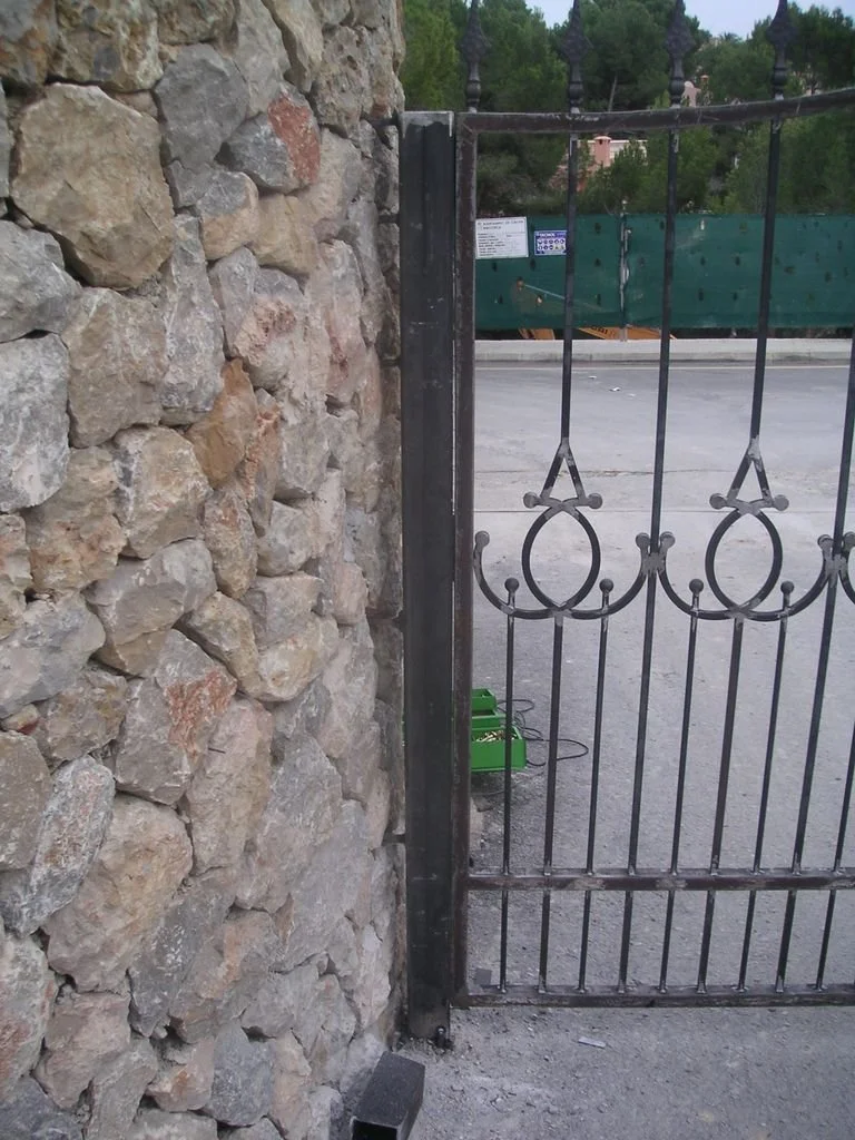 Puerta de hierro con muro de piedra al lado, en una calle con árboles y cartel en el fondo.