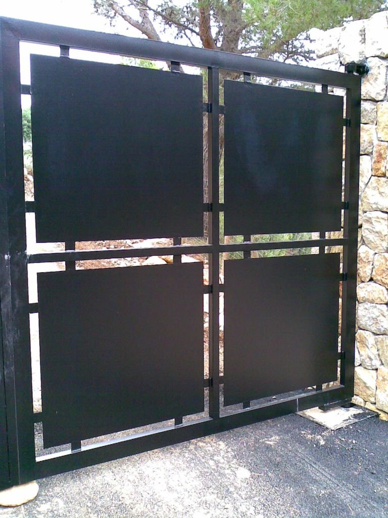 Puerta de metal negra con diseño geométrico y piedras en la pared al fondo, en un entorno exterior.