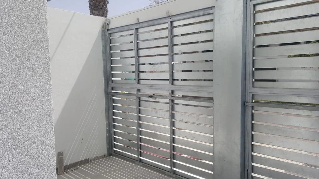Puerta metálica de listones horizontales en un patio con paredes blancas y sombra en un día soleado.
