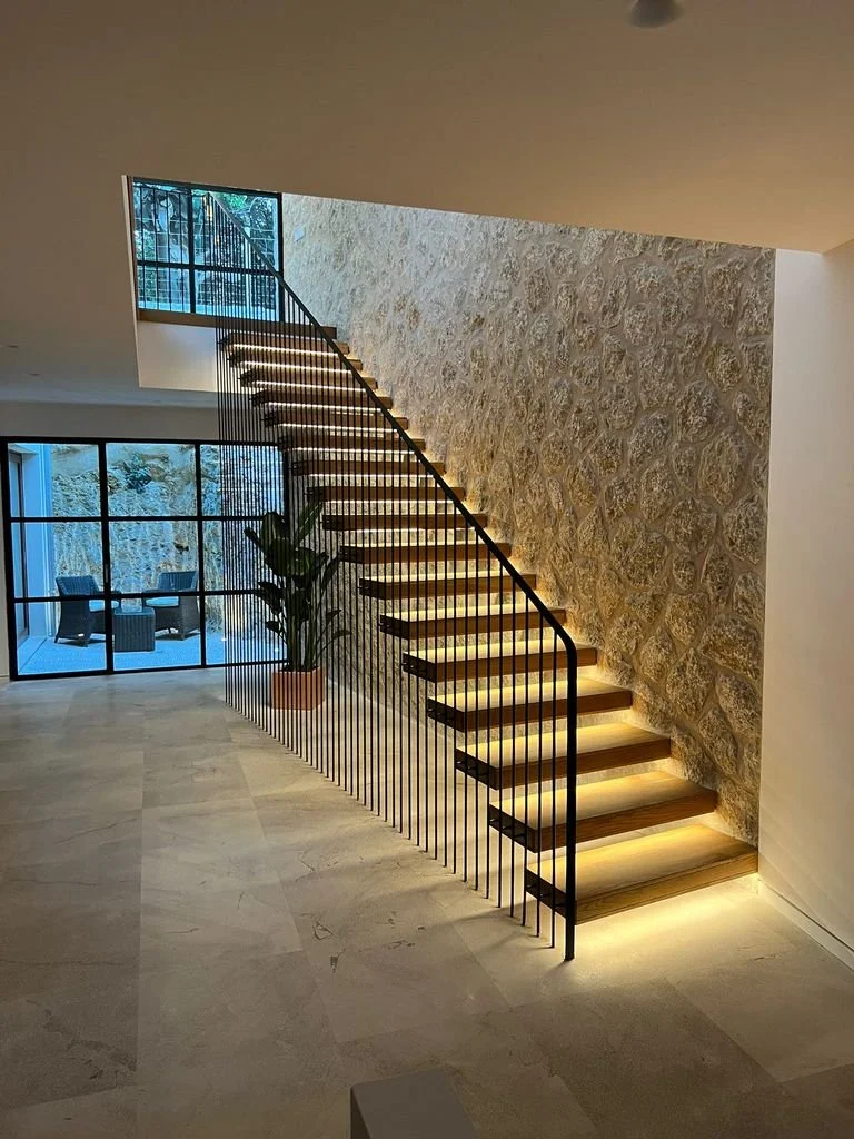 Escalera moderna de madera con iluminación empotrada, una pared de piedra y ventanas grandes en una casa elegante.