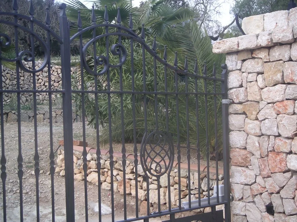 Puerta de hierro forjado negra con un muro de piedra a la derecha y un jardín con plantas verdes atrás.