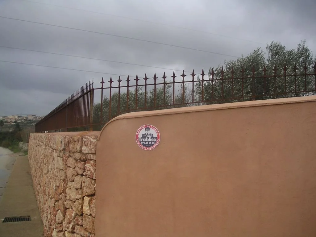 Valla con muro de piedra y reja de metal en un día nublado, con un sticker en la pared que dice 'Trabisa' y un teléfono.