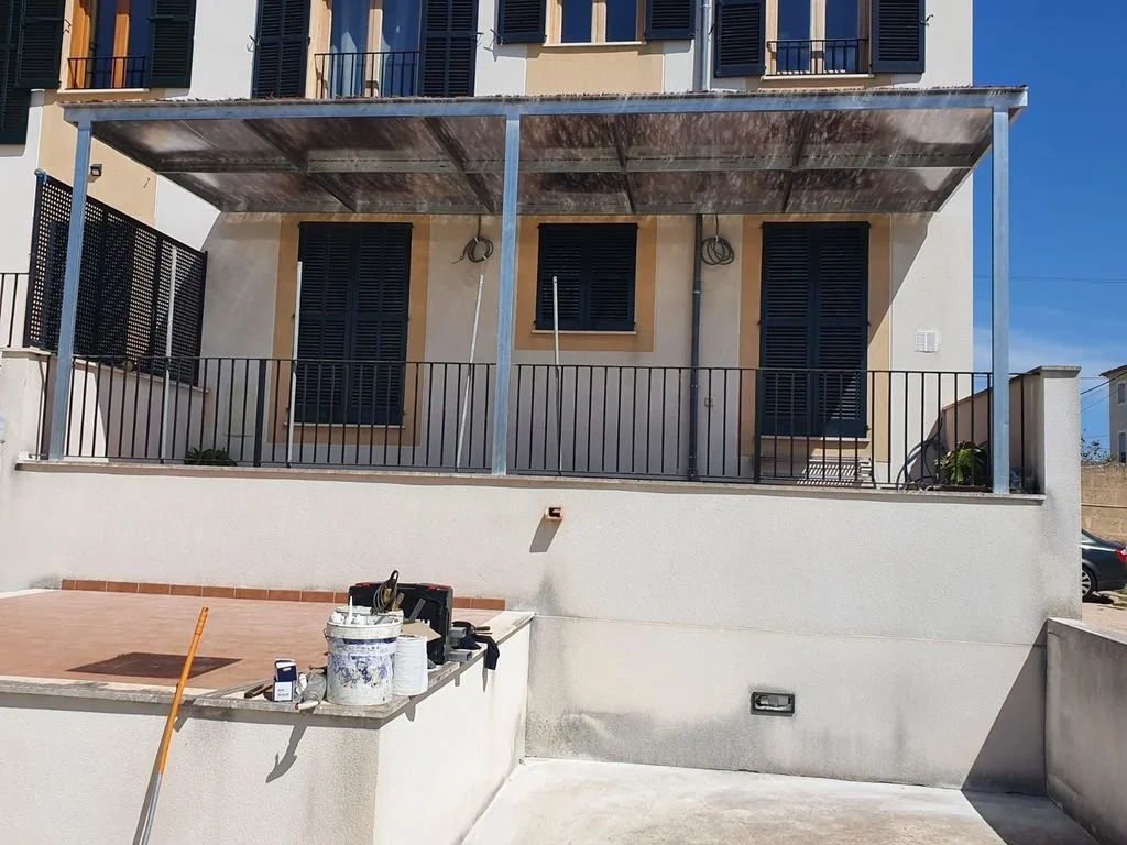 Fachada de una casa con balcón y estructura metálica para un toldo. En la parte inferior, hay materiales de construcción y herramientas en una superficie de trabajo.