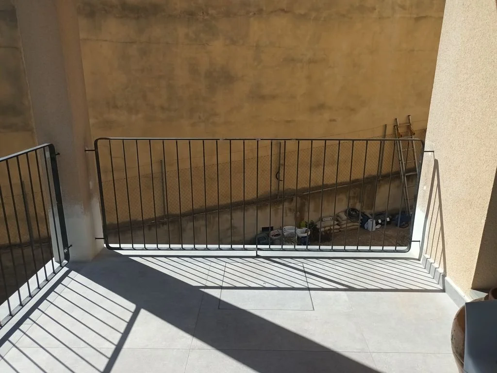 Balconera de metal con sombra proyectada en el piso, vista a un área de tierra con herramientas y una escalera