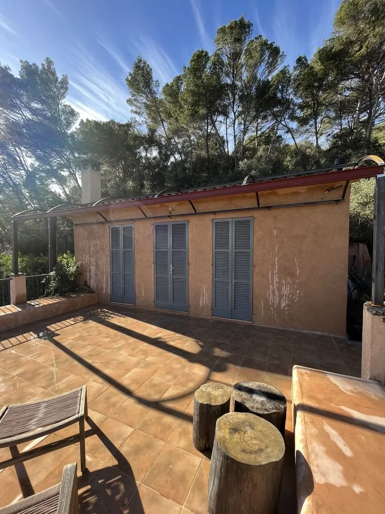 Terraza con suelo de baldosas, muebles de madera y vista a un edificio con paredes de color naranja y puertas de persiana gris, rodeada de árboles.