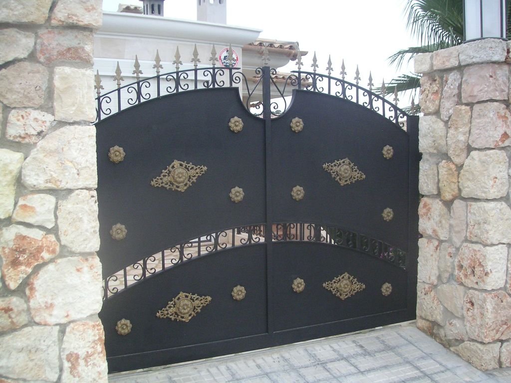 Puerta negra de hierro con decoraciones doradas, entre columnas de piedra y con un jardín visible al fondo.