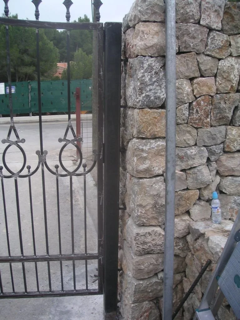 Una cerca de hierro con adorno en la parte superior y una columna de piedra a su lado, en un ambiente externo, con un bote de agua en una roca cercana.