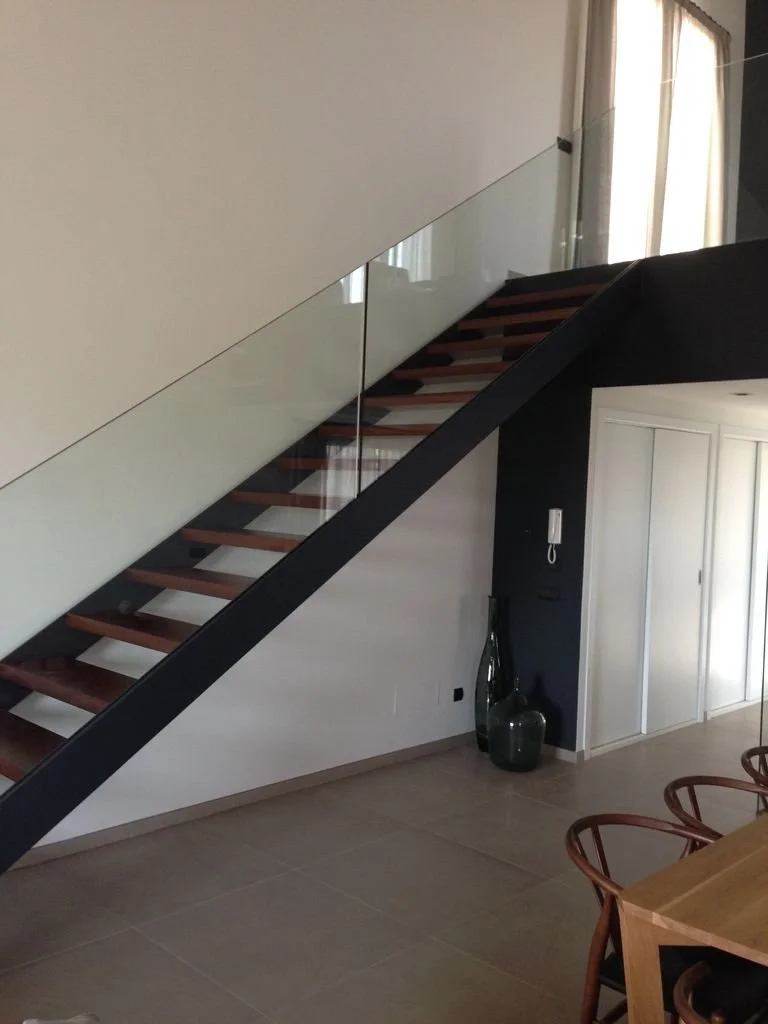 Escalera moderna con barandilla de vidrio en un salón interior, con decoración minimalista y ventanas que dejan entrar luz natural.