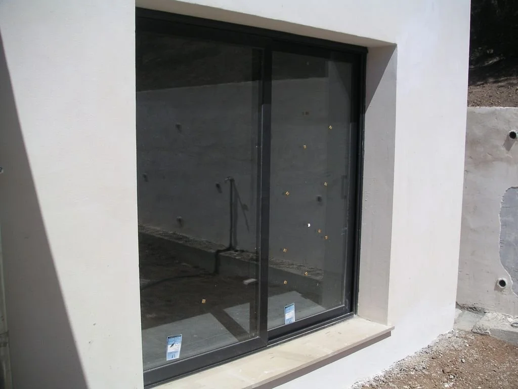 Ventana de vidrio con marco negro en una pared blanca, en construcción, con tierra y material de construcción alrededor.
